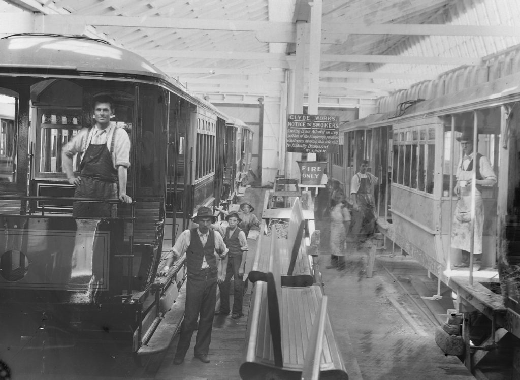 Black and white photograph of two trams parked inside a large carriage workshop surrounded by a group of men. Some are wearing work aprons with others in vests and trousers. A sign on a pole in the background reads ‘CLYDE WORKS’. 